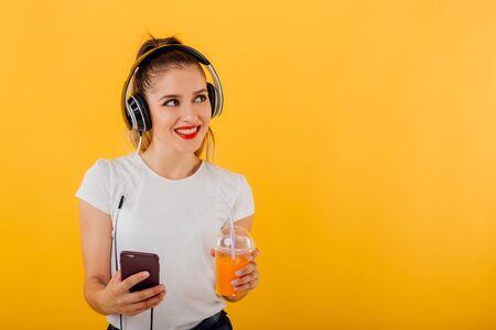 nice girl smiles listening to music, in the smartphone hand has headphones head, with juice cup, dressed in a white shirt,  positive facial emotion isolated on a yellow background, copy spaceの写真素材