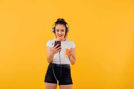 nice girl smiles talking on the phone, has headphones on head, with juice cup, dressed in a white shirt,  positive facial emotion isolated on a yellow background, copy spaceの写真素材