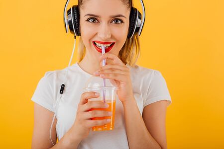 cropped. smiling girl looks at the camera, is the head headphones,  plastic cup of juice dressed in a white shirt,  positive facial emotion isolated on a yellow background, copy spaceの写真素材