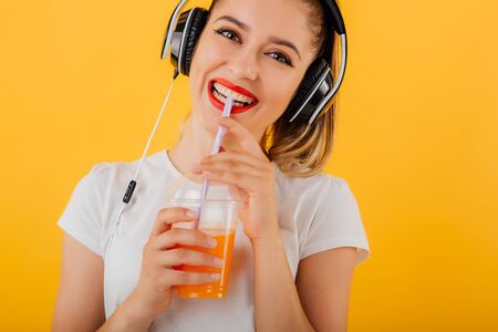 Close up. smiling girl looks at the camera, is the head headphones, drink juice cup dressed in a white shirt,  positive facial emotion isolated on a yellow background, copy spaceの写真素材
