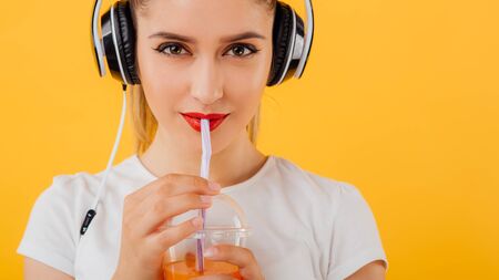 Close up. red lips beautiful young girl, look at the camera, is holding plastic cup with juice dressed in a white shirt,  positive facial emotion isolated on a yellow backgroundの写真素材