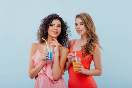fashionable two young girls with curly hair, hold the plastic cup with juice, look at the camera positive facial emotions, isolated on blue background, copy spaceの写真素材