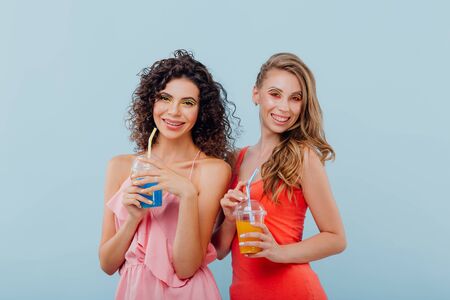 fashionable two cheerful girls with curly hair, hold the plastic cup with juice, look at the camera positive facial emotions, isolated on blue background, copy spaceの写真素材