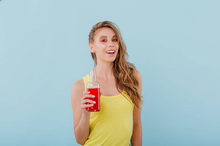 beautiful young girl smiles having the bottle in her hand and looks into the camera, positive facial emotions, dressed in yellow shirt isolated on blue background, copy spaceの写真素材