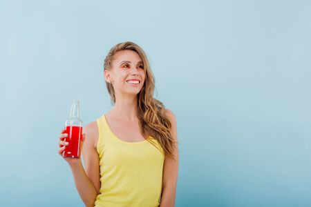 young girl smiles amazefully, has the drink bottle in her hand and looks sideways, positive facial emotions, idressed in yellow shirt solated on blue background, copy spaceの写真素材