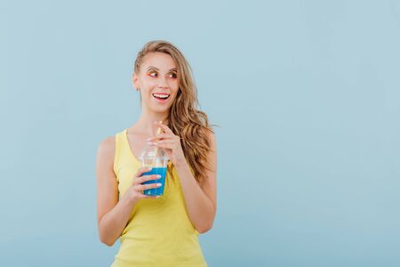 beautiful girl smiles amazed, has the plastic cup with the drink in his hand and looks sideways, dressed in yellow shirt, positive facial emotions, isolated on blue background, copy spaceの写真素材