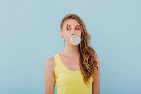 beautiful girl chewing gum bubbles, dressed in yellow shirt, positive facial emotions, isolated on blue background, copy spaceの写真素材