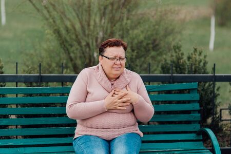 Middle-age lady touching her chest, suffering from heart attack, disease, dressed in a pink sweater and in jeans sitting on a chair in the parkの写真素材