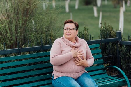 overweight Woman feeling sharp pain in her chest, suffering heart attack , health , dressed in a pink sweater and in jeans sitting on a chair in the parkの写真素材