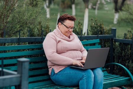 overweight middle-aged woman using laptop, dressed in a pink sweater and in jeans sitting on a chair in the parkの写真素材