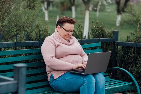 overweight middle-aged woman uses laptop, she is happy, dressed in a pink sweater and in jeans sitting on a chair in the parkの写真素材