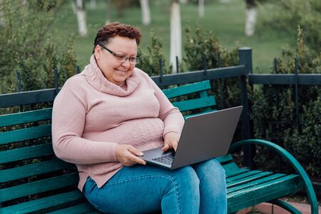 middle-aged woman uses the laptop, dressed in a pink sweater and in jeans sitting on a chair in the parkの写真素材