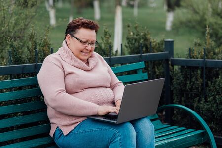 middle-aged woman in glasses uses her laptop, she is happy, dressed in a pink sweater and in jeans sitting on a chair in the parkの写真素材