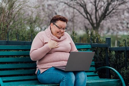 middle-aged woman in glasses is surprised uses notebook is happy, dressed in a pink sweater and in jeans sitting on a chair in the parkの写真素材