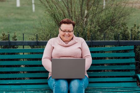 middle-aged woman in glasses using laptop messaging with friend sharing funny videos and news communicating online, dressed in a pink sweater and in jeans sitting on a chair in the parkの写真素材