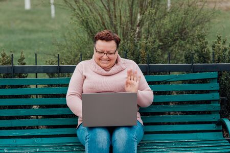 Alone middle aged mature woman using laptop messaging with friend sharing funny videos and news communicating online, dressed in a pink sweater and in jeans sitting on a chair in the parkの写真素材