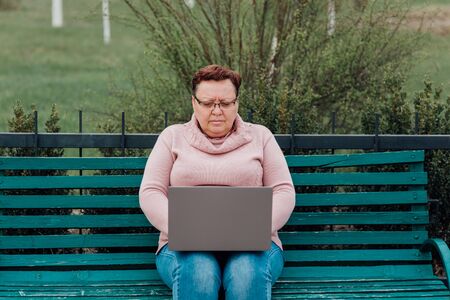woman sweeping in glasses using the notebook, is worried, dressed in a pink sweater and in jeans sitting on a chair in the parkの写真素材