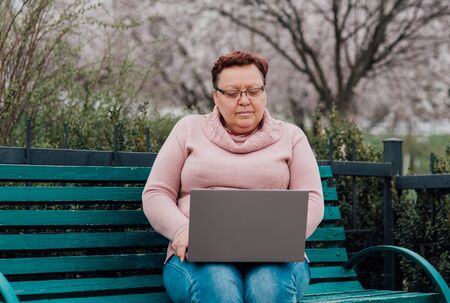 woman sweeping in glasses using notebook, dressed in a pink sweater and in jeans sitting on a chair in the parkの写真素材
