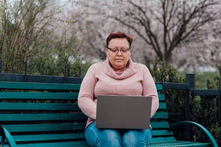 middle-aged woman in glasses uses the notebook, dressed in a pink sweater and in jeans sitting on a chair in the parkの写真素材