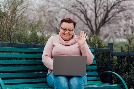 Alone middle aged mature woman using laptop messaging with friend sharing funny videos and news communicating online, dressed in a pink sweater and in jeans sitting on a chair in the parkの写真素材