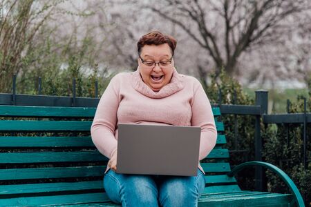 middle-aged woman in glasses is surprised using notebook is happy, dressed in a pink sweater and in jeans sitting on a chair in the parkの写真素材