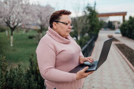 middle-aged woman in glasses uses laptop, makes shopping online, dressed in a pink sweater and in jeans, outdoorsの写真素材