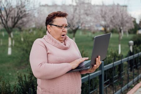 middle-aged woman in glasses using laptop, is scared, dressed in a pink sweater and in jeans, outdoorsの写真素材