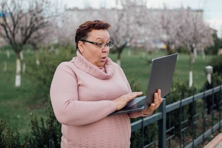 mature woman in glasses using laptop, is scared, dressed in a pink sweater and in jeans, outdoorsの写真素材