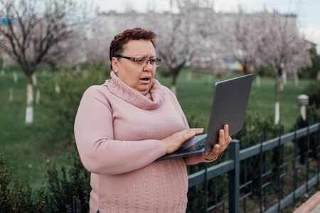 woman sweeping in glasses uses her laptop, she's scared, she saw what she did not need to see, dressed in a pink sweater and in jeans, outdoorsの写真素材