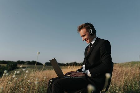 young happy businessman with headphones using laptop, in field, sitting on chair, outdoorの写真素材