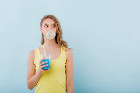 young girl chewing gum bubbles, has plastic cup with juice in hand, dressed in yellow shirt, positive facial emotions, isolated on blue background, copy spaceの写真素材