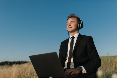 young man with headphones listening music in field, sitting on chair, using laptop, eyes closed,...