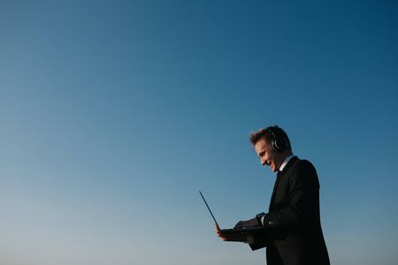 excited young businessman using laptop, has headphones, copy space, profile view, sky background, outdoorの写真素材