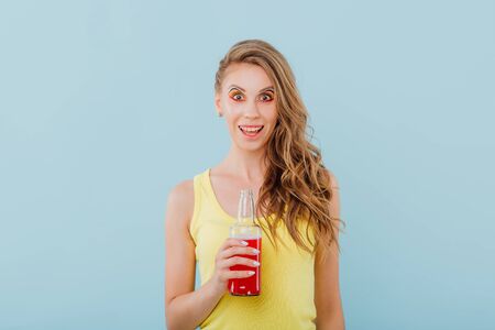 young girl smiles amazed, has the drink bottle in her hand and looks into the camera, positive facial emotions, dressed in yellow shirt isolated on blue background, copy spaceの写真素材