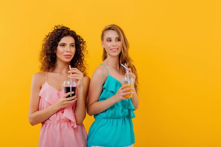 two cheerful women with curly hair, holding a plastic cup with juice in a pink and blue shirt, positive facial emotions, isolated on yellow background, copy spaceの写真素材