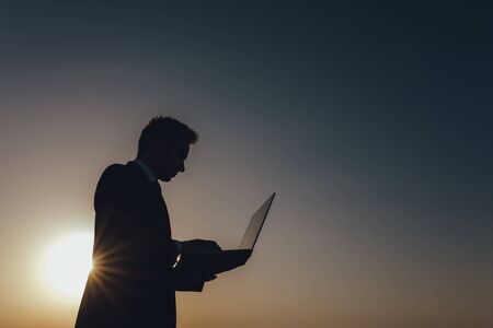 young businessman using laptop, Internet and social network concept, at sunset, copy space, profile view, sky background, outdoorの写真素材