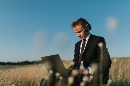 young businessman with headphones using video chatting, in field, sitting on chair, outdoorの写真素材