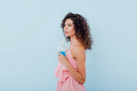 beautiful young lady with curly hair dressed in pink shirt, holding plastic cup with juice, isolated on blue background, copy spaceの写真素材