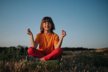 little girl deals with yoga, dressed in orange shirt, at sunset, outdoorsの写真素材