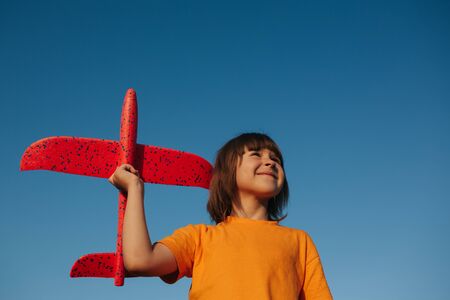 Beautiful kid holding in hands a red toy airplane on a background of blue sky, outdoors, sky background, copy spaceの写真素材