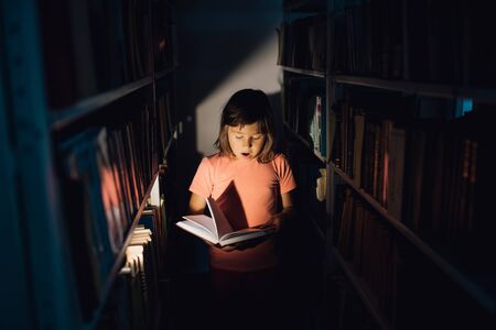 little girl reads the book, is scared, has open mouth. holds a book in his hand, learns in the library, indoorsの写真素材