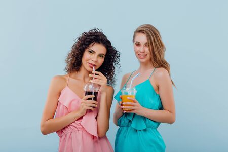 two happy women with curly hair smile and hold the plastic cup with juice in pink and blue shirt, positive facial emotions, isolated on blue background, copy spaceの写真素材