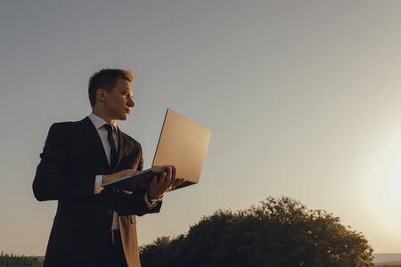 young businessman using laptop, Internet and social network concept, at sunset, copy space, profile view, sky background, outdoorの写真素材