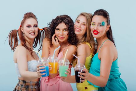 four happy young girls with unusual hairstyle and make-up, holding plastic juice cups, looking at the camera isolated blue background, positive facial emotions, copy spaceの写真素材