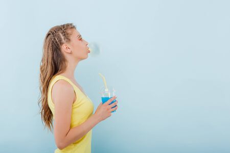 close up. young girl chewing gum bubbles, has plastic cup with juice in hand, dressed in yellow shirt, side view, positive facial emotions, isolated on blue background, copy spaceの写真素材
