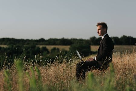 Young businessman on meadow,  sitting in the chair, using a laptop, outdoor, profile viewの写真素材