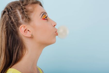 close up. young girl chewing gum bubbles, dressed in yellow shirt, side view, positive facial emotions, isolated on blue background, copy spaceの写真素材
