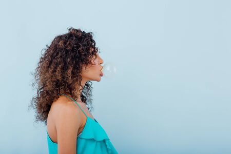 fashionable young girl chewing gum bubbles, with curly hair, dressed in blue shirt, isolated on blue background, copy spaceの写真素材