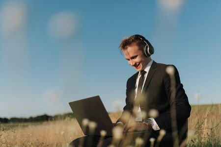 young happy businessman with headphones using video chatting, in field, sitting on chair, outdoorの写真素材