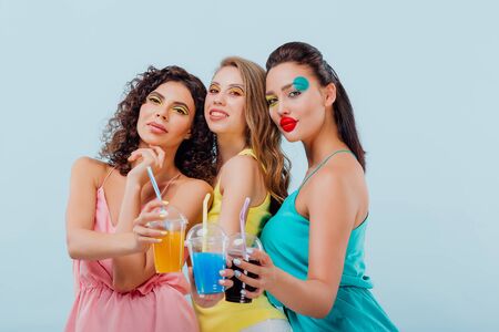 three girls girlfriends happy with hairstyle and makeup unusual, is holding plastic cups of juice, look at the camera, isolated blue background, positive facial emotions, copy spaceの写真素材
